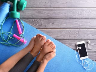 A woman stretching her legs on a blue yoga mat with her phone, weights and a jump rope