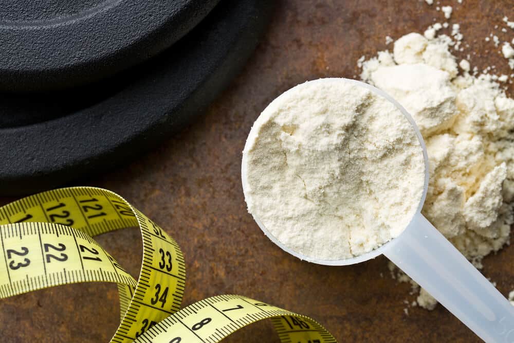A scoop of white whey protein on a brown counter with yellow and black measuring tape and a black weight.
